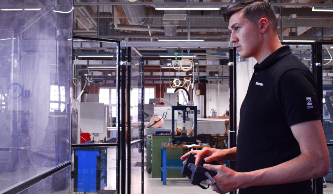 En elev står med en robotkontroller i handen. Mittemot eleven är det en plastvägg, Bakom eleven är ett verkstadsutrymme.